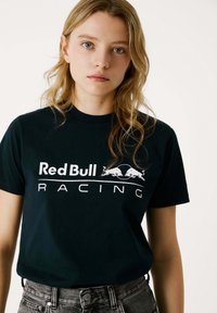 Melnas kokvilnas kreklas ar "Red Bull Racing" logo baltā krāsā, īsām piedurknēm, apaļā kakla izgriezuma un brīvā piegriezumā. Zem krekla redzami džinsa šorti.