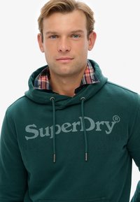Zielona bluza z kapturem, z sznurkami, z dużym szarym logo "SuperDry". Wyłożony kaptur ma wielokolorowy wzór w kratę. Tkanina mieszanka bawełny.