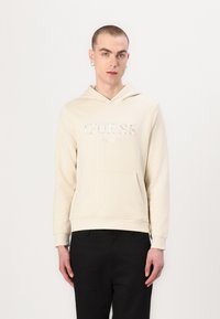 Sudadera color crema con un bolsillo delantero y capucha, con un logotipo plateado bordado "GUESS EST. 1981" en el pecho. Textura suave.