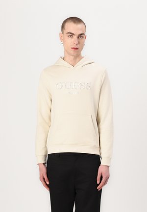 Kremfarget hettegensere med lomme foran og hette, med en sølvbrodert "GUESS EST. 1981" logo på brystet. Myk tekstur.