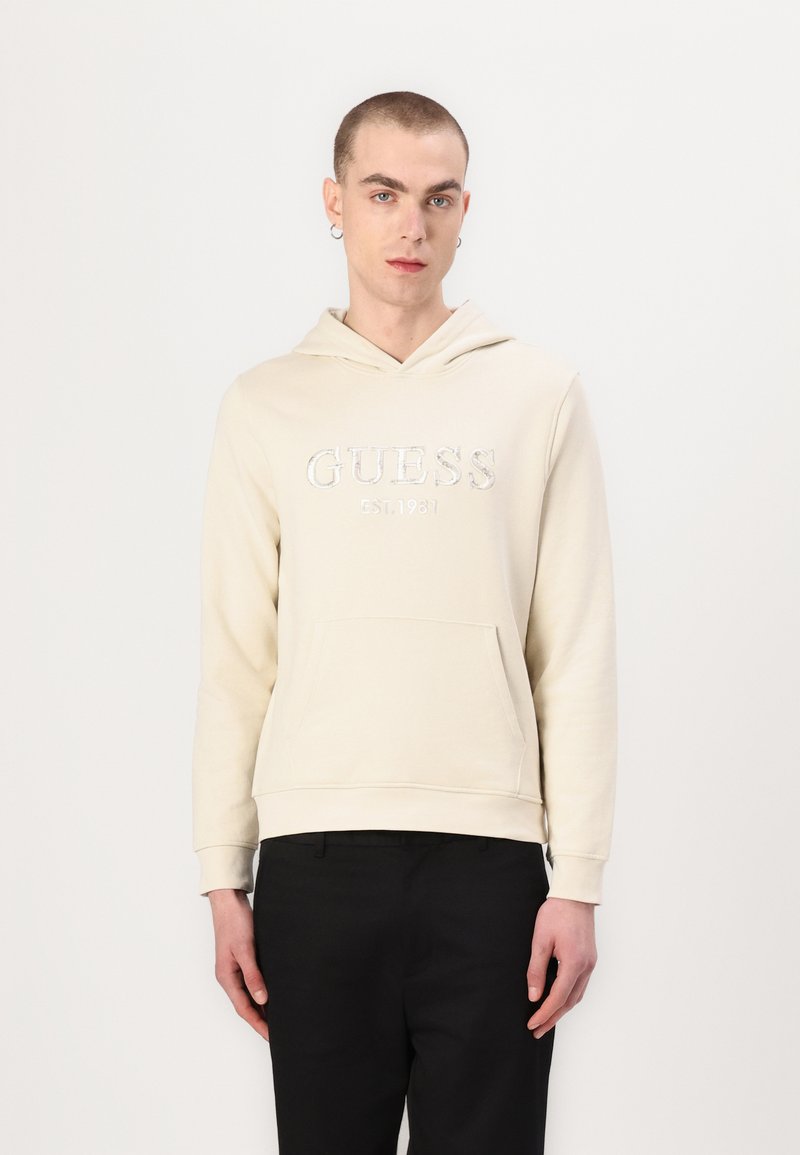 Sudadera color crema con un bolsillo delantero y capucha, con un logotipo plateado bordado "GUESS EST. 1981" en el pecho. Textura suave.