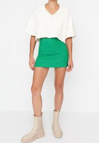 Chemise blanche raccourcie avec deux poches avant, associée à une mini-jupe verte texturée et des bottines beiges à semelles épaisses.