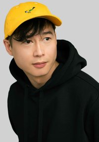 Żółta czapka z wyszytym ptakiem, noszona z czarnym kapturem. Miękki materiał, zakrzywiony daszek i regulowany pasek z tyłu.