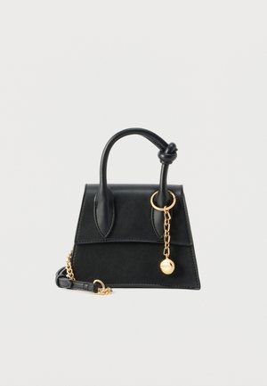 Petit sac à main trapézoïdal noir avec une anse nouée, une sangle chaîne dorée, et un charme doré pendu à l'anse.