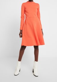 Långärmad, figurnära korallklänning med hög krage, utsvängd kjol och slät textur, i kombination med glansiga vita ankelskor.