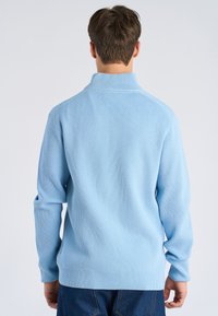 Uomo di spalle che indossa un maglione a coste azzurro e jeans blu scuro su uno sfondo semplice.