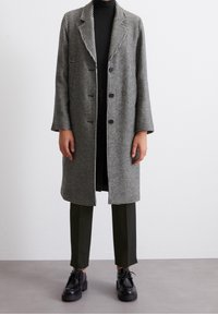 Abrigo de pata de gallo en gris y negro con cuello con muescas, botones de doble pecho y mangas largas, combinado con pantalones oscuros y zapatos.