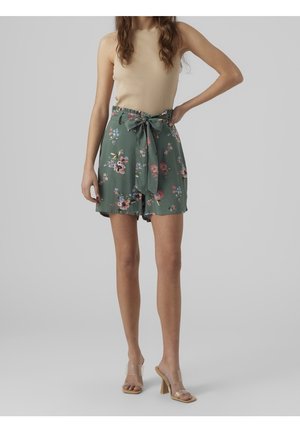 Shorts floraux verts avec un détail de taille à nouer, fabriqués en tissu léger. Associés à un débardeur beige en maille côtelée et des sandales à talons transparents.