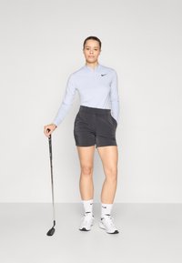 Maglia da golf azzurro chiaro a maniche lunghe con zip a metà, pantaloni corti neri, calzini bianchi e scarpe da ginnastica bianche, che tiene una mazza da golf. Tessuto morbido.