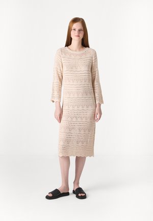Robe en tricot beige à manches trois quarts, présentant un motif géométrique en dentelle et un ourlet festonné ; assortie avec des sandales noires à enfiler.