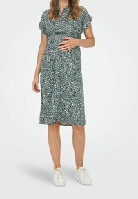 Robe de maternité en sarcelle avec un imprimé feuille, manches courtes, patte de boutonnage et coupe décontractée. Associée à des baskets blanches.