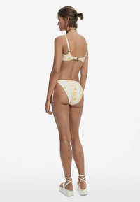 Bikini-set in het wit met een geel aquarel patroon, met een sluiting op de rug, verstelbare bandjes en hooggesneden onderstukken met minimale bedekking.