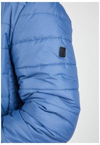 Blauer Puffermantelärmel mit gestepptem Muster, ausgestattet mit einem kleinen schwarzen Etikett am Arm. Das Material scheint leicht und isoliert zu sein.