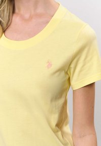 Kollane puuvillane T-särk ümar kaelusega, lühikeste varrukatega ja väike roosa tikandiga logo rinnal. Sile tekstuur ja liibuv lõige.