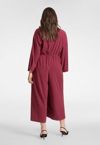 Bordowy kombinezon o szerokich nogawkach i długich rękawach, z paskiem w talii modelującym sylwetkę. Gładka faktura, noszony z czarnymi szpilkami.