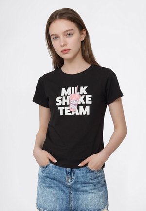 Млада жена с кафява коса, носеща черна тениска с надпис "MILKSHAKE TEAM" и розова графика на млечен шейк, и синя дънкова пола.