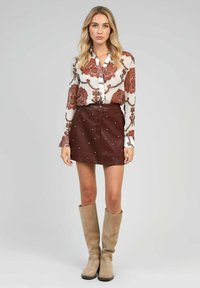 Chemise blanche à manches longues avec un motif floral orange, associée à une mini jupe en cuir bordeaux ornée de clous en argent, et des bottes beiges jusqu'aux genoux.
