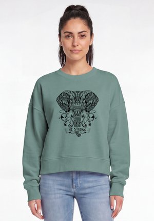 Vrouw met donker haar in een staart draagt een teal sweatshirt met een gedetailleerd zwart olifantenontwerp en de tekst "Be Strong" op de voorkant.