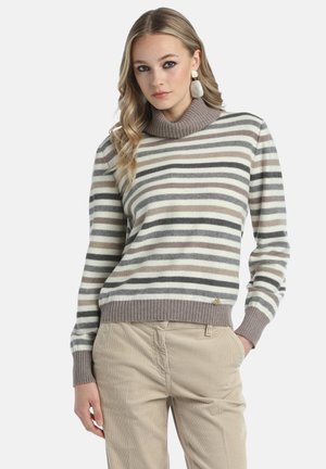 Striped sweater in cream, beige, and gray with a high ribbed collar and ribbed cuffs. Metal logo detail near the hem. Paired with beige corduroy pants.