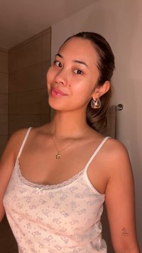 Camisola bianca leggermente decorata con pizzo; collana dorata e grandi orecchini a cerchio in argento. Pelle liscia, dettagli dello sfondo minimi.