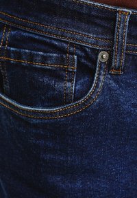 Ciemnoniebieskie dżinsy z kontrastowymi pomarańczowymi szwami, z przednią kieszenią i metalowym zapięciem. Faktura wydaje się gładka.