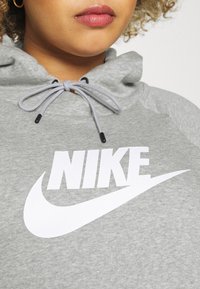 Grå hoodie med en stor vit Nike-logga, med en justerbar huva med snoddar som har svarta spetsar. Texturerat tyg.