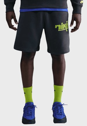 Persona che indossa pantaloncini neri Nike con logo giallo, calzini Nike giallo acceso e scarpe da ginnastica Nike blu, in piedi con le mani rilassate ai lati.