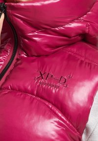 Polstrovaná bunda jasně růžové nylonové barvy, s vysokým límcem a zipem. Vyšité logo "XP-D+" a "Superdry" na látce.