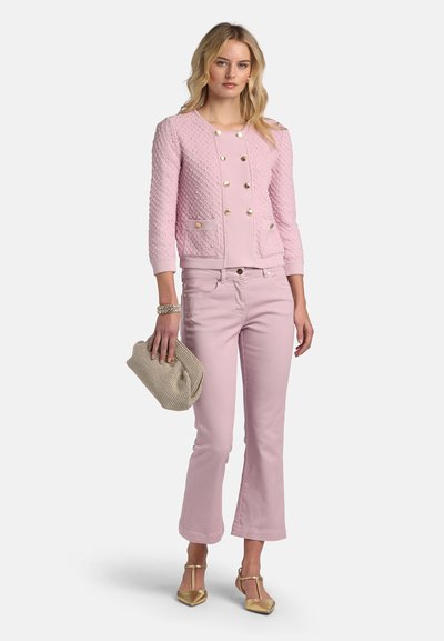 Femme portant une veste rose texturée avec des boutons dorés, un pantalon rose assorti, des talons dorés, tenant une pochette beige tissée.