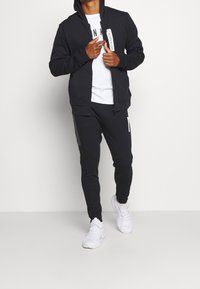 Haut à capuche zippé noir avec capuche et étiquette de logo, associé à un jogger sombre avec des accents latéraux et des baskets blanches. Texture de tissu lisse.