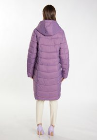 Kvinde står med ryggen til kameraet iført en mellemlang quiltet lilla jakke med hætte, cremefarvede bukser og lyslilla højhælede sko.