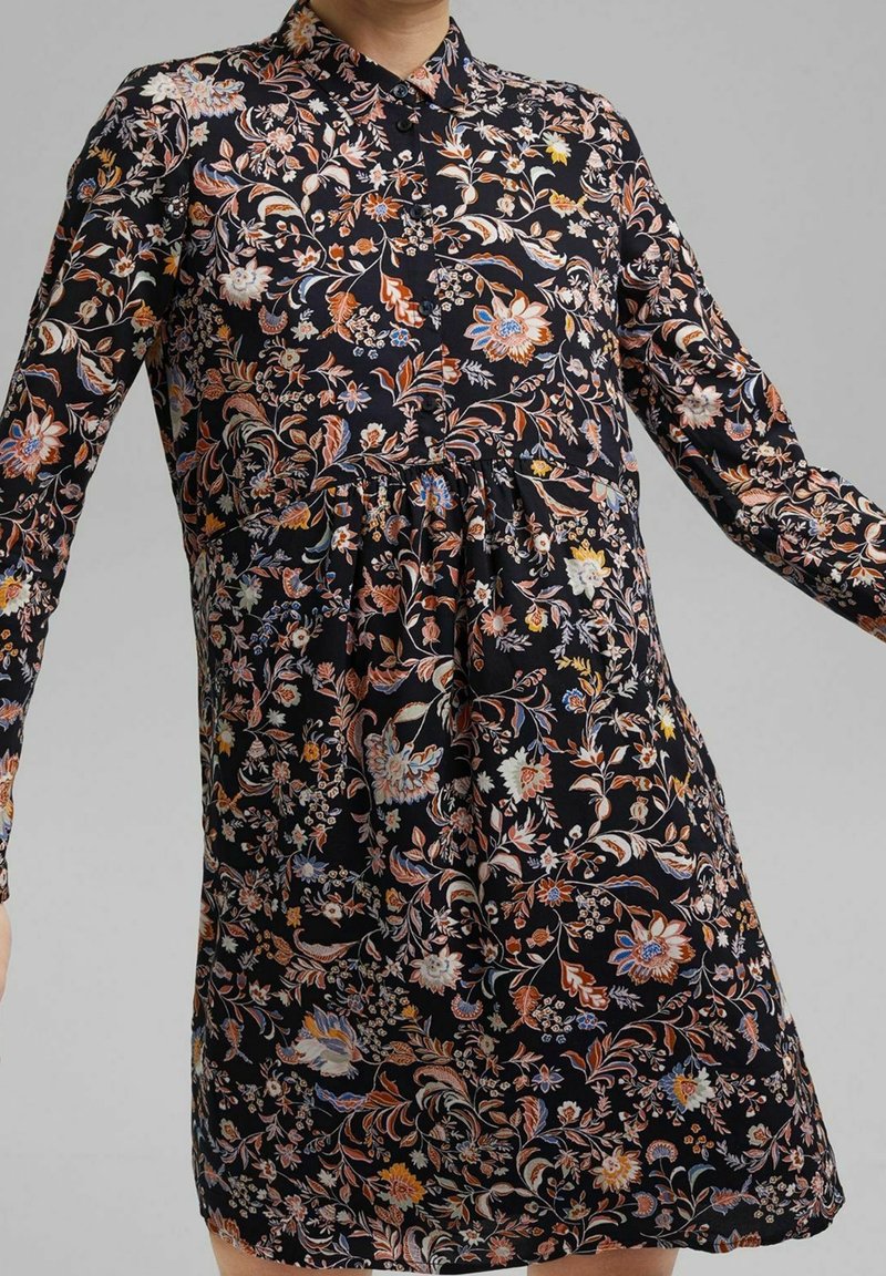 Blommig klänning i svart tyg med långa ärmar, krage och samlad midja. Mönster inkluderar färgglada blommor och lövverk.