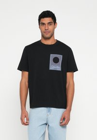 Melna kokvilnas t-krekls ar taisnstūrveida grafiku violetā un baltā krāsā ar horizontālām līnijām un centrālu apli. Īsas piedurknes, apaļš kakls.