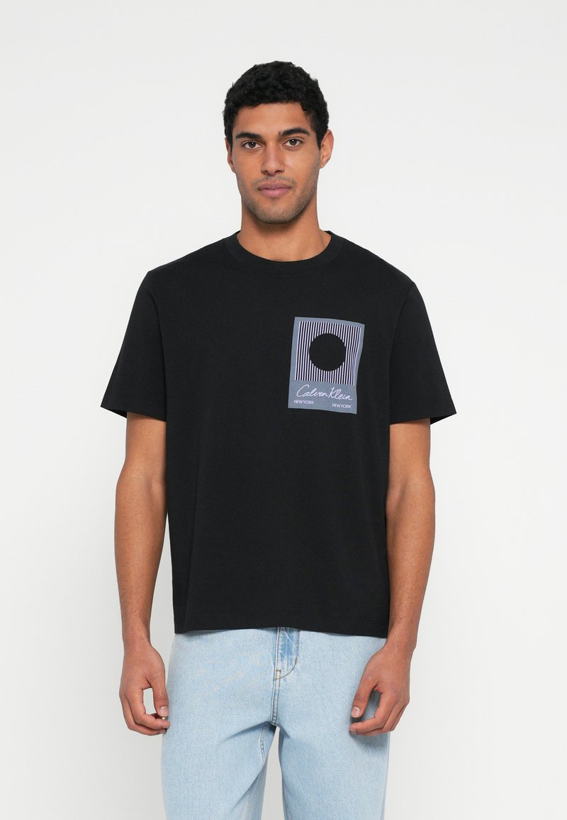 Melna kokvilnas t-krekls ar taisnstūrveida grafiku violetā un baltā krāsā ar horizontālām līnijām un centrālu apli. Īsas piedurknes, apaļš kakls.