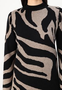 El suéter de punto presenta un patrón de rayas de cebra en negro y beige, con un escote redondo y mangas largas, mostrando una tela suave y texturizada.