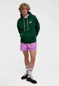 Green hoodie with white drawstrings and front logo, paired with pink shorts, black shoes, and striped socks. Casual athletic attire.