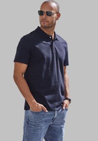 Navyblaues Poloshirt mit klassischem Kragen und kurzen Ärmeln, kombiniert mit hellblauen Jeans. Das Modell trägt dunkle Sonnenbrillen und hat eine Uhr an.