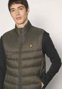 Olive zaļš pūkains vests ar augstu apkakli un rāvējslēdzēju. Iezīmējas ar horizontālu šuvju zīmējumu un mazu dzeltenu logotipu uz krūtīm.