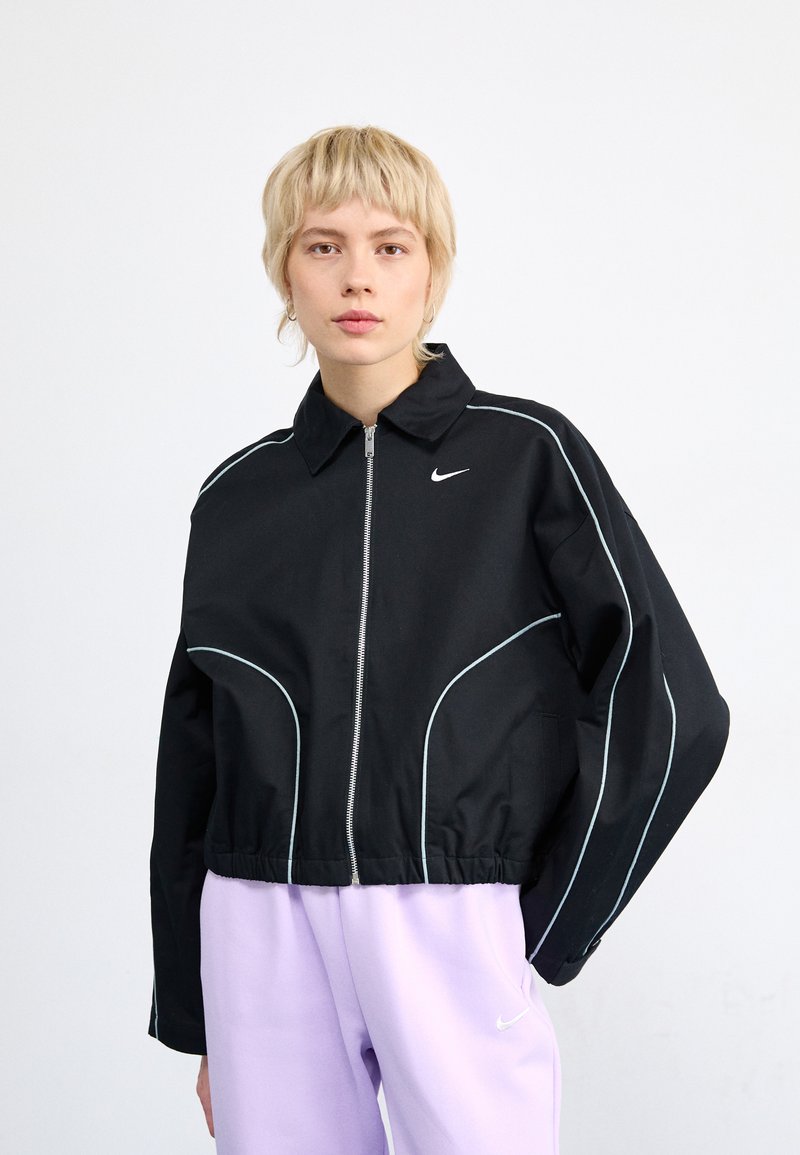 Chaqueta negra corta con cremallera frontal, diseño de ribete blanco y cuello; logo de Nike en el lado izquierdo del pecho. Combinada con pantalones de chándal lavanda.