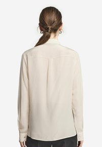 Ljust beige långärmad blus med en slät textur, en klassisk krage och en enda bakfåll. Har knäppta manschetter och en rundad nederkant.