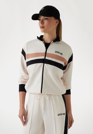 Giacca bianca con zip e strisce nere e pesca, con il logo "GUESS 1981". Abbinata a pantaloni coordinati, entrambi in tessuto morbido. Un berretto nero adorna la testa.