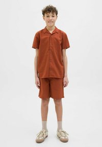 Roestkleurige set van een kortekorte mouwen shirt en shorts, met een gestructureerde stof. Gecombineerd met lichtbeige sneakers en geribbelde sokken.