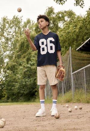Navy sports jersey with large white "86," beige shorts, white sneakers with blue stripes. Child holding a baseball glove, surrounded by baseballs.