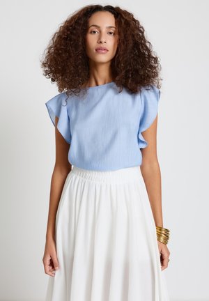 Frau mit lockigem Haar, die ein hellblaues Oberteil mit Rüschenärmeln, einen weißen Rock und mehrere goldene Armreife am Handgelenk trägt.