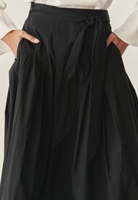 Zwarte geplooide rok met een strikceintuur, gemaakt van een gestructureerde stof, met zijzakken en een vloeiende silhouet. Gedetailleerd met zachte plooien.
