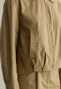 Chaqueta corta de tela suave. Presenta un cierre frontal, un cuello de punta y un bolsillo lateral. Mangas ligeramente abullonadas.