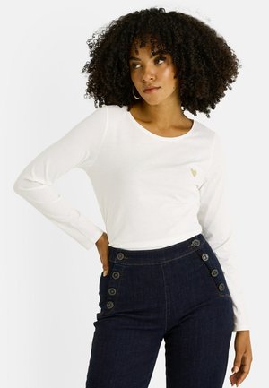 Top blanco de manga larga con cuello redondo y pequeño detalle de corazón dorado; combinado con pantalones de mezclilla azul oscuro de cintura alta con detalles de botones.