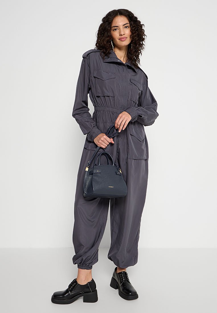 Tuta grigio leggero con maniche lunghe, cintura elastica e due tasche frontali. Scarpe nere chunky con lacci e una piccola borsa a tracolla blu navy.