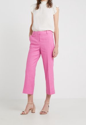 Vrouw draagt een wit mouwloos blouse met ruches, felroze cropped broek en beige sandalen met hakken tegen een effen achtergrond.