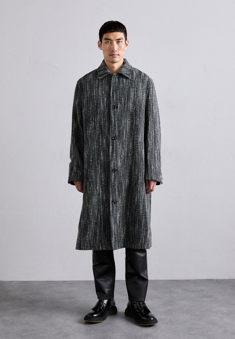 Manteau long, gris et noir à motifs, en tissu texturé, présentant une fermeture à boutonnage, un grand col et une coupe légèrement oversize.
